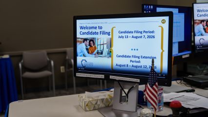 A computer monitor shows information about the candidate filing period from July 13 to August 7, 2026, and the extension period from August 8 to August 12, 2026.