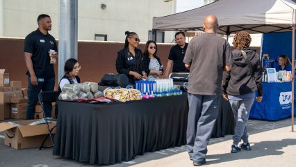 Five individuals from the Office of Homeless Services are standing and seated at a table with supplies as attendees walk by for information and giveaways.