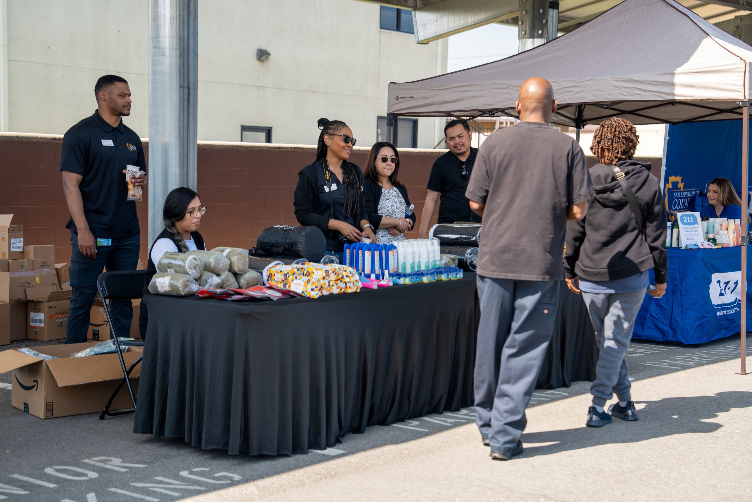 Five individuals from the Office of Homeless Services are standing and seated at a table with supplies as attendees walk by for information and giveaways.