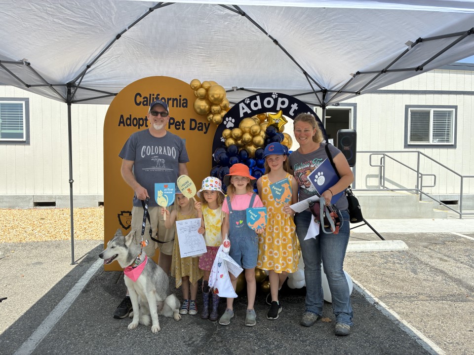 Community members pose with their newly adopted pet in front of a California Adopt-A-Pet Day backdrop decorated with balloons.b