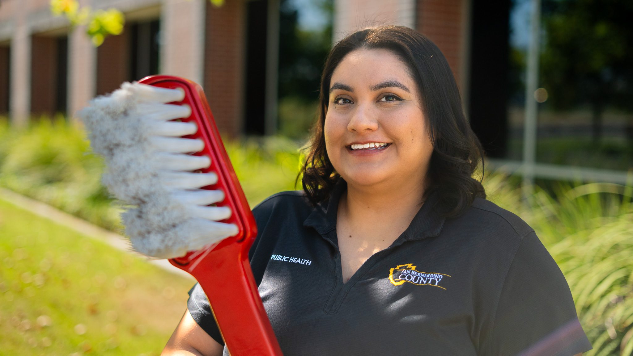 Woman smiling outdoors holding an oversized red toothbrush.