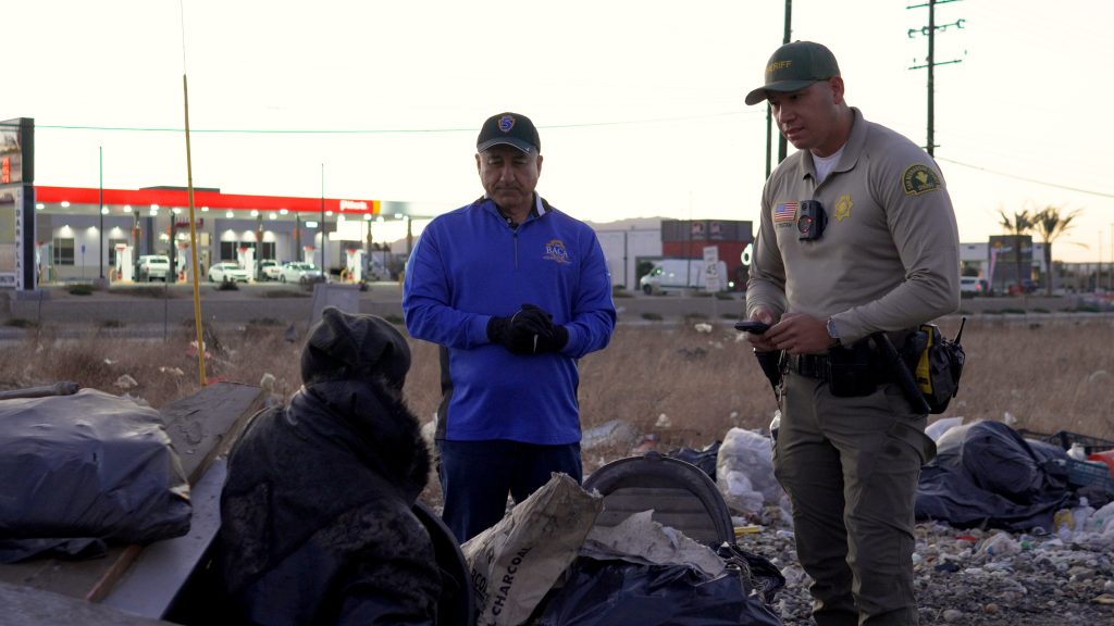 Two individuals, one in a San Bernardino County Sheriff’s uniform and another in a royal blue jacket, stand outside and engage with a homeless individual.