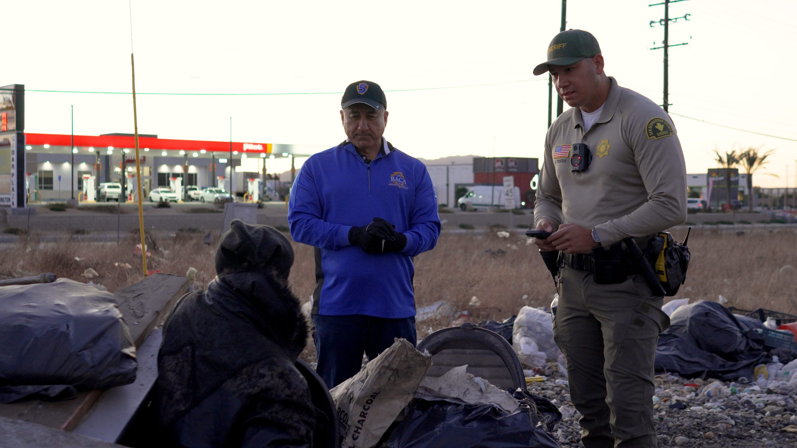 Two individuals, one in a San Bernardino County Sheriff’s uniform and another in a royal blue jacket, stand outside and engage with a homeless individual.