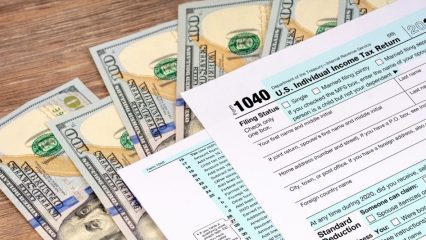 A tax form labeled '1040 U.S. Individual Income Tax Return' sits on a wooden table, with a spread of $100 bills visible underneath.