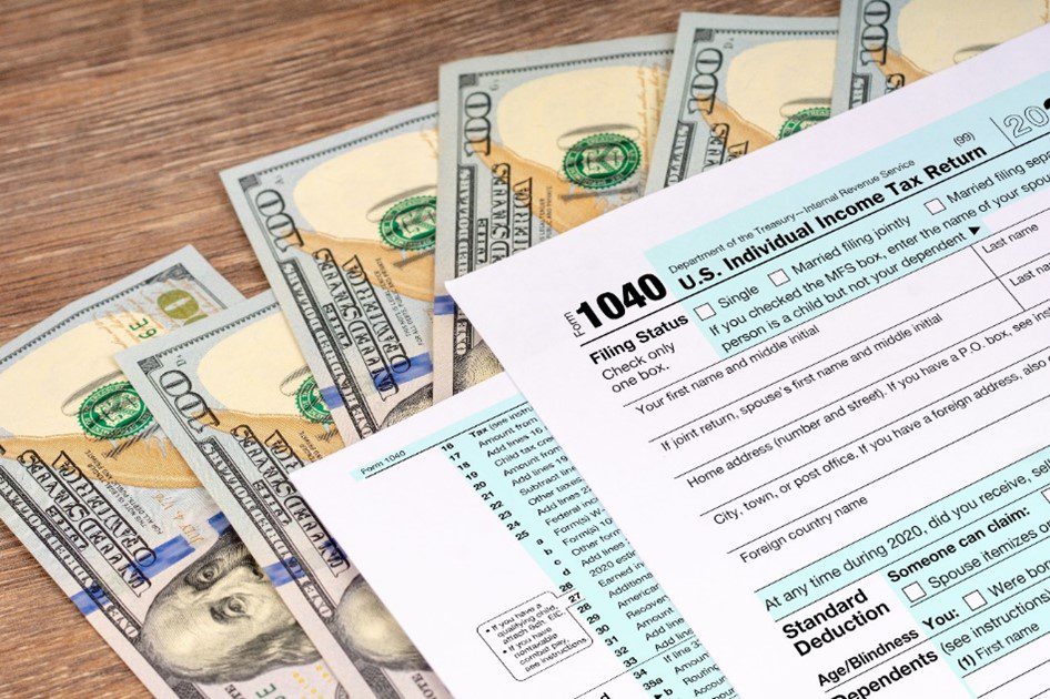 A tax form labeled '1040 U.S. Individual Income Tax Return' sits on a wooden table, with a spread of $100 bills visible underneath.