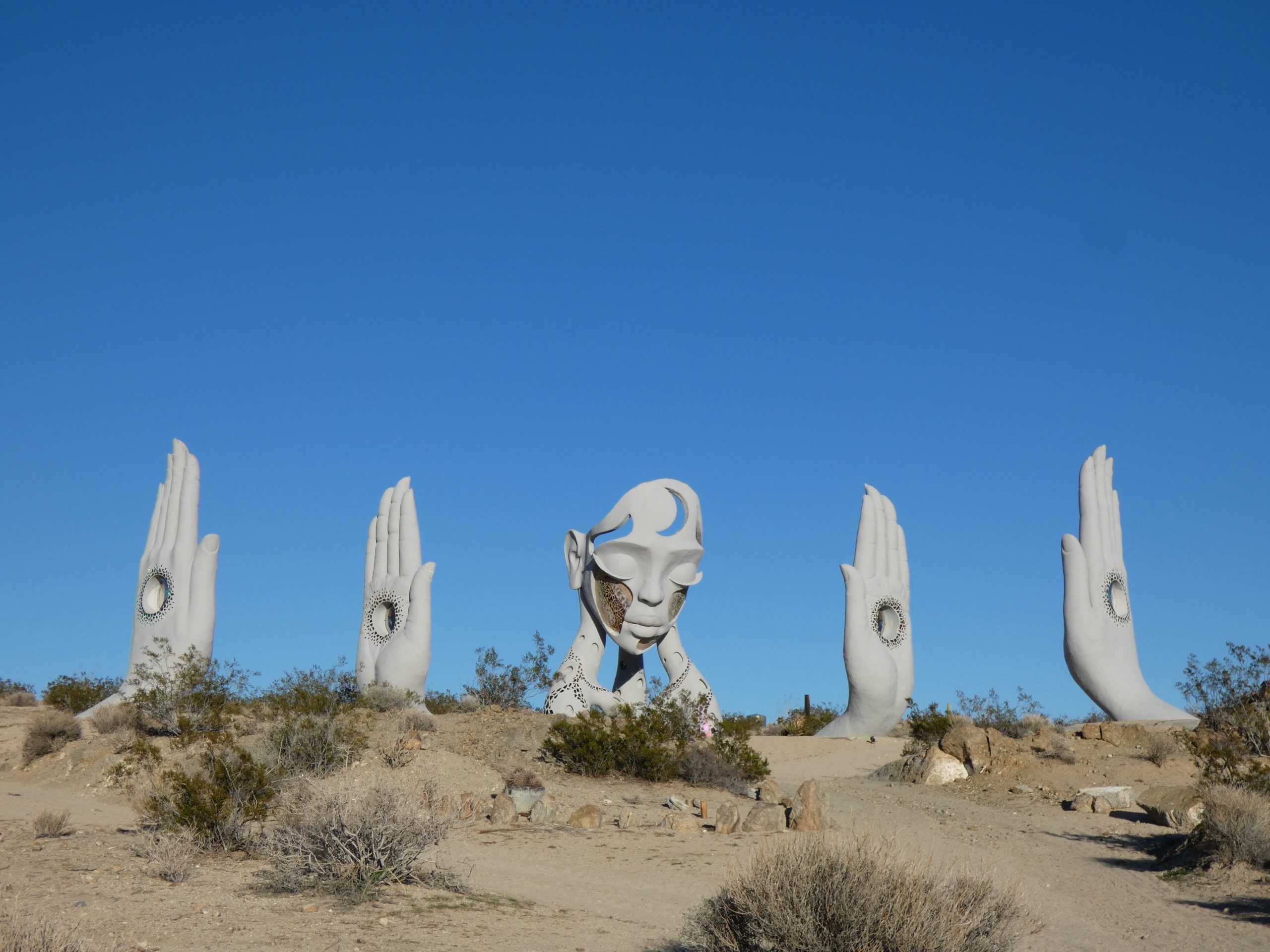 A desert landscape features a large art installation with a face and four hands rising from the ground.