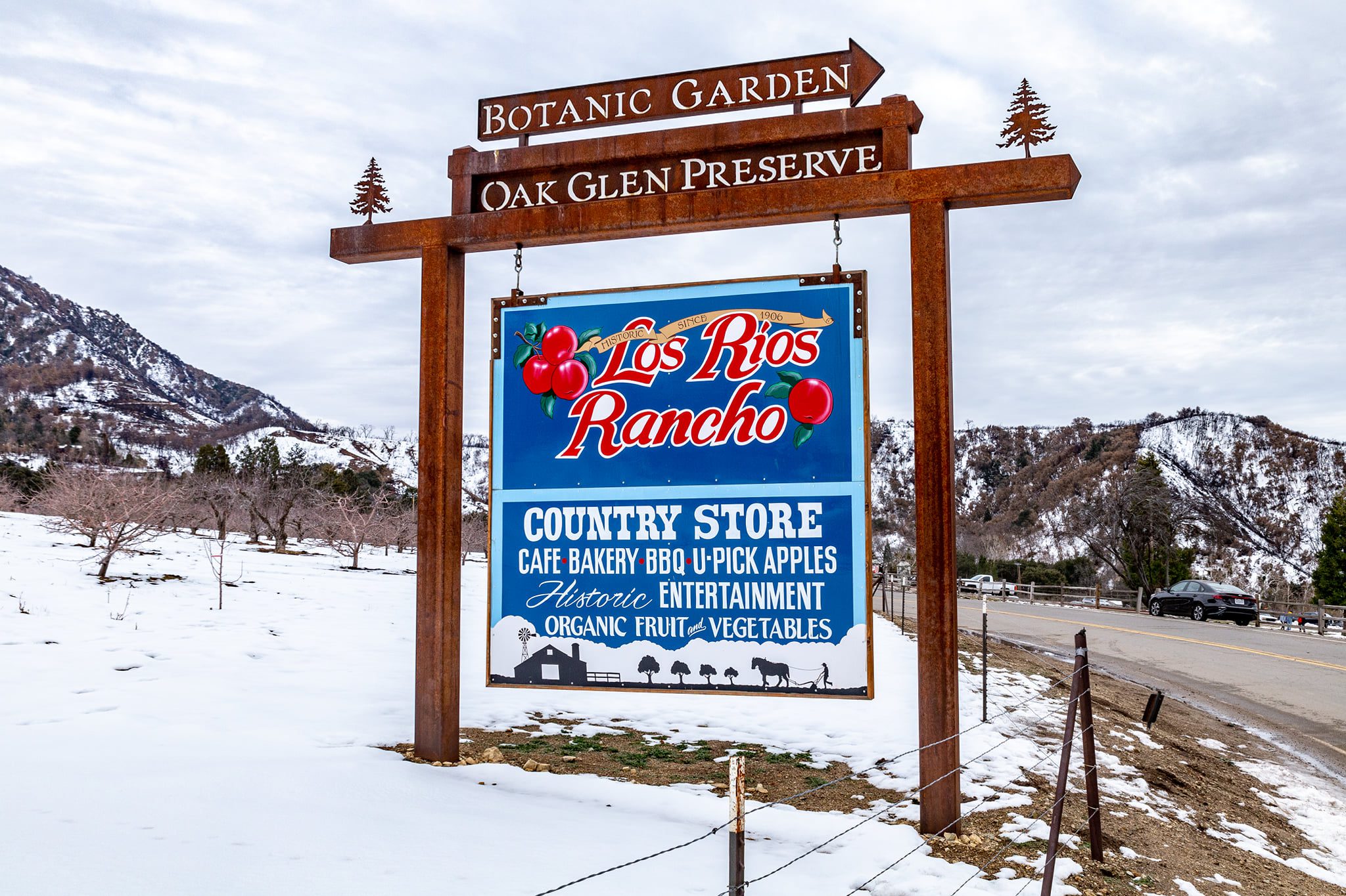 A rustic entrance sign for Los Rios Rancho in Oak Glen, featuring red apples and text promoting a country store, U-pick apples, a cafe, bakery, and organic produce. The sign is mounted on a metal frame with directional signs for the Oak Glen Preserve and Botanic Garden, set against a snow-covered orchard with mountains in the distance and a winding road.