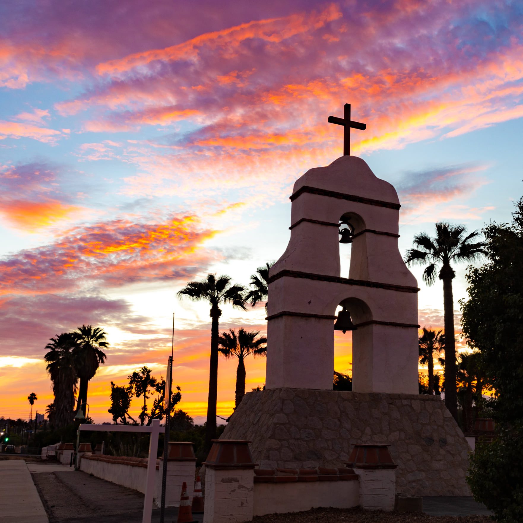An image of a bell tower with a cross on top taken at sunset featuring palm trees in the background.
