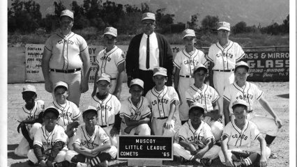 A black and white photo of a baseball team with players and a coach. The team is named "Muscoy Little League Comets," as indicated by a sign in front of the group.