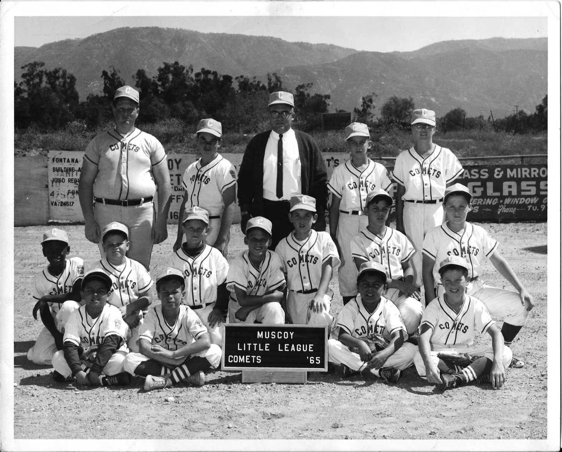 A black and white photo of a baseball team with players and a coach. The team is named "Muscoy Little League Comets," as indicated by a sign in front of the group.