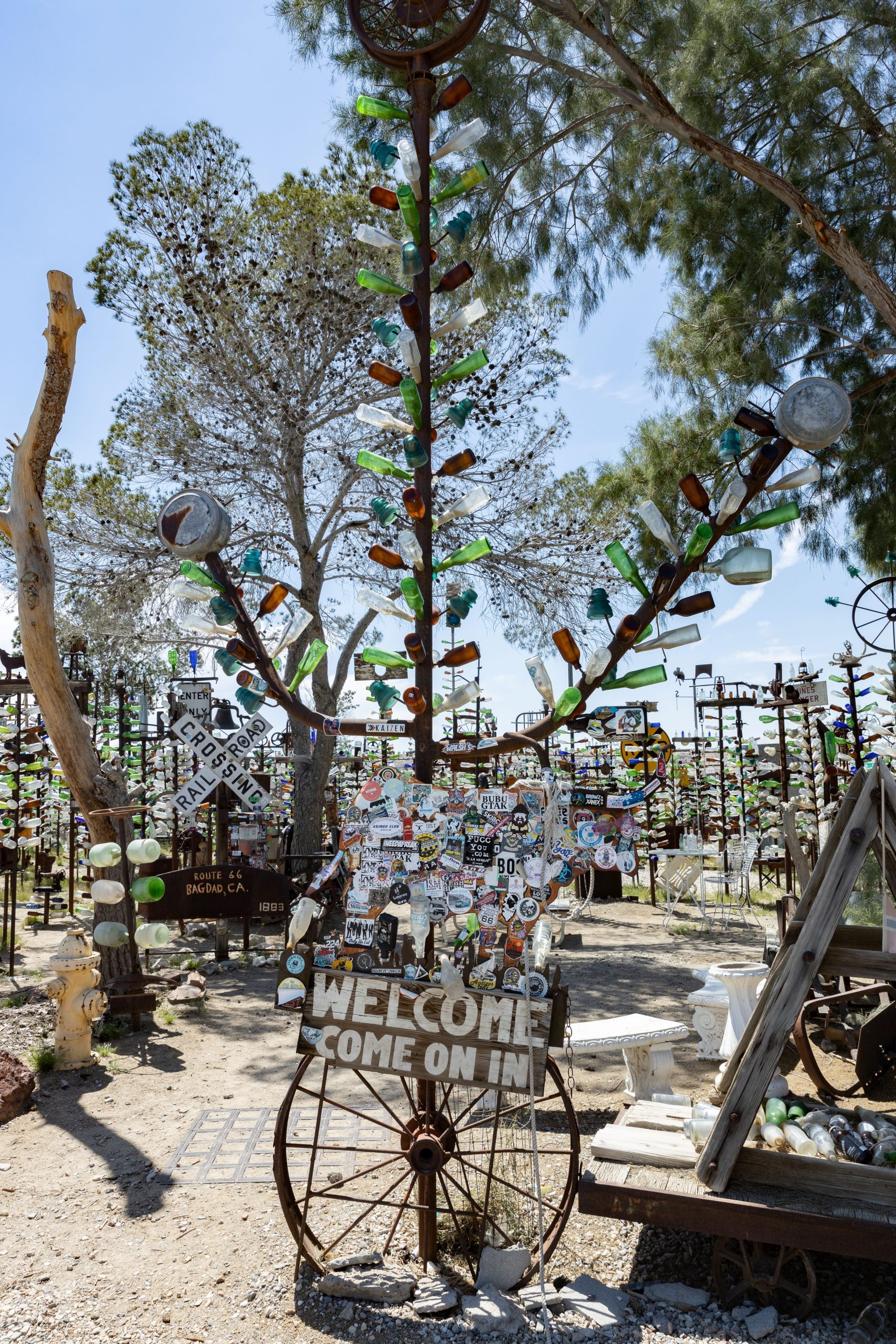 Colorful glass bottles arranged on old metal pipes and tree limbs with a sign that says “Welcome, come on in.”