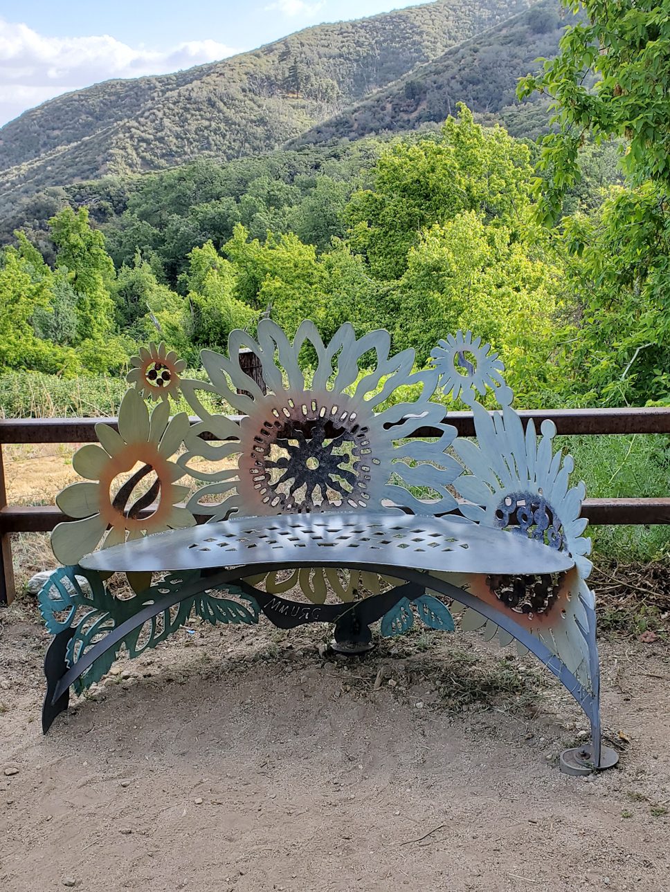 A metal bench with sunflower designs is positioned on a dirt path and surrounded by tall trees.