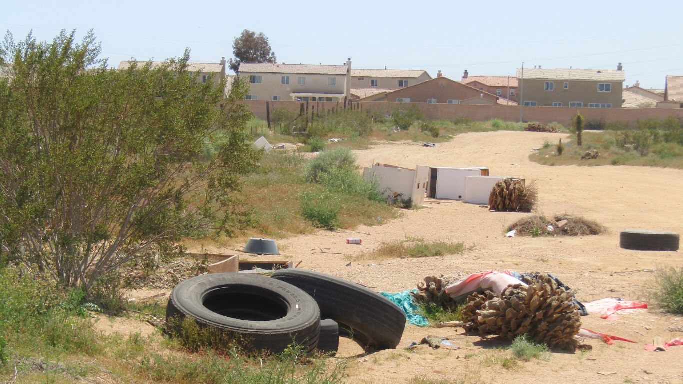 Old tires and discarded trash lie in a field lining the desert field with a track of homes seen in the background.