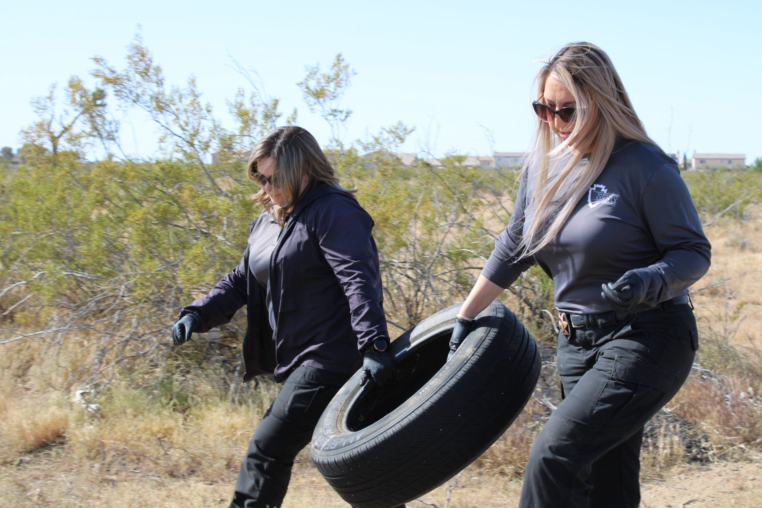 Two female code enforcement officers lift and carry a tire walking on a dirt road in the desert.