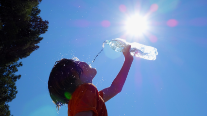 A young boy sprays himself with water drops from a water bottle he is holding with the sun overhead and a tree to the left and the blue sky.