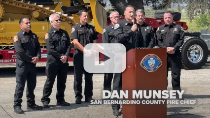 A group of County firefighters stand at a podium in front of a bulldozer with a video play button.
