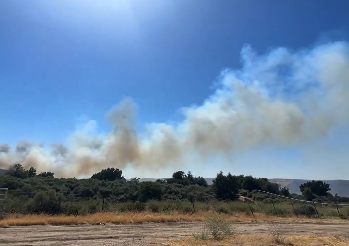 A photo of a fire with a big plume of smoke in the desert against a blue sky with trees and brush along a dirt road.