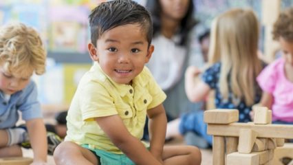 An Asian boy toddler looks into the camera smiling sitting inside a classroom setting.