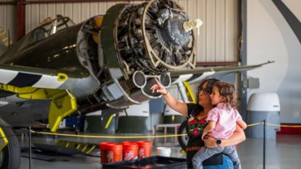 A woman holding a young girl in her arms point in the air with a vintage plane behind her in a airport hangar.