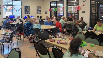 A lively senior center with tables and chairs filled with people socializing. Colorful banners and decorations add a festive atmosphere to the bright room.