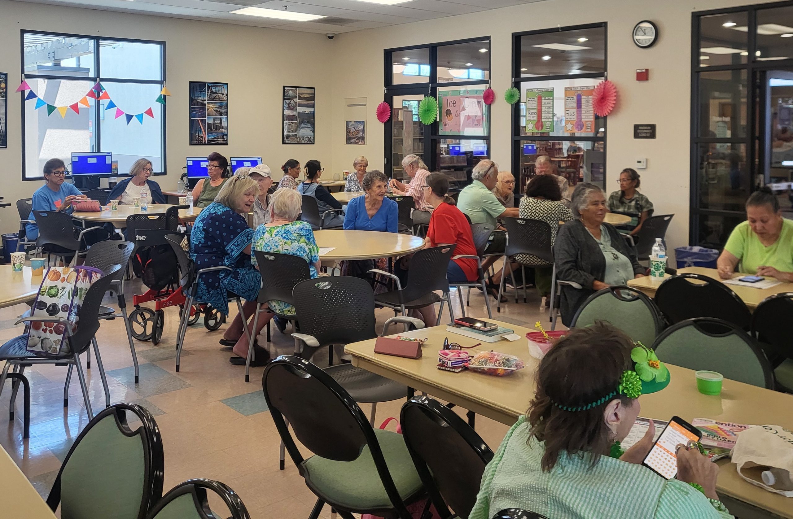 A lively senior center with tables and chairs filled with people socializing. Colorful banners and decorations add a festive atmosphere to the bright room.