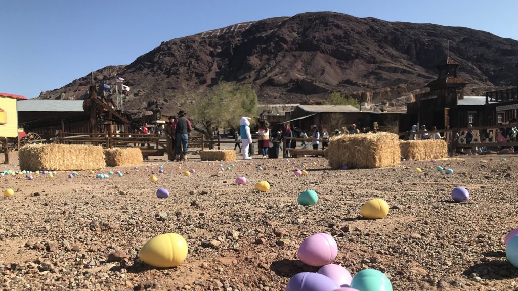 Plastic Easter eggs on a dirt ground with the Easter bunny, people and hay bales in the background at Calico Ghost Town.