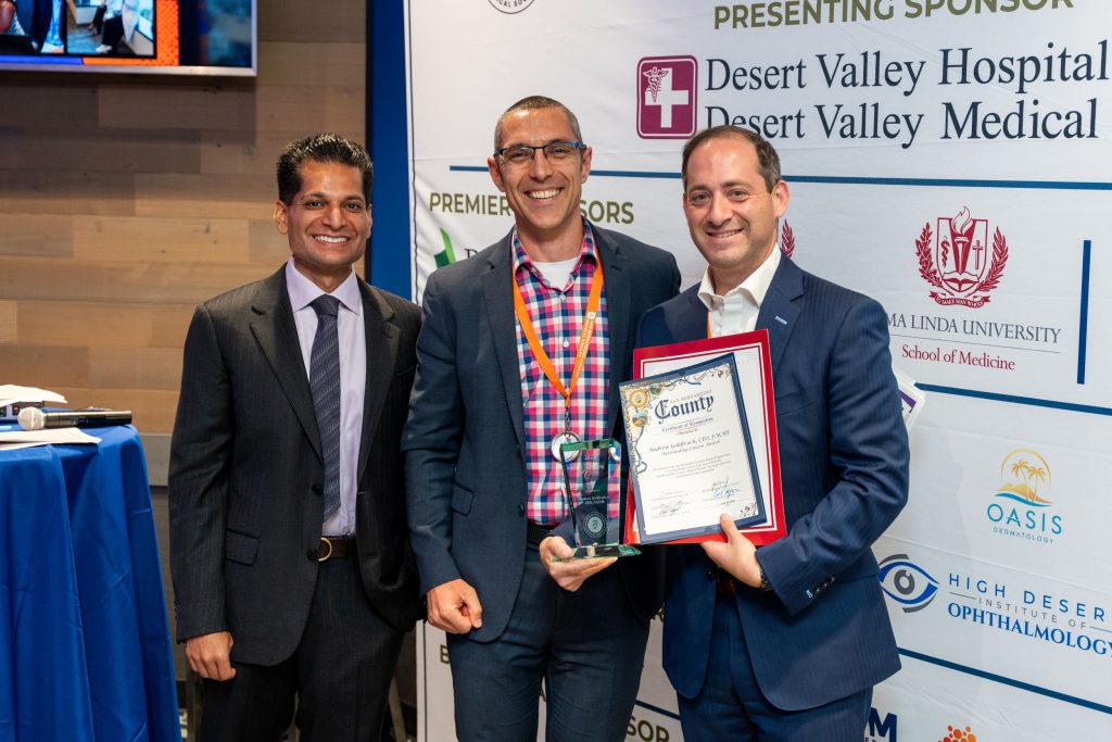 Three individuals are standing together, smiling. One person on the right is holding an award and certificate. The backdrop displays logos for Desert Valley Hospital, Loma Linda University School of Medicine, and other sponsors.