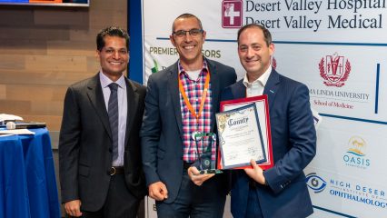 Three individuals are standing together, smiling. One person on the right is holding an award and certificate. The backdrop displays logos for Desert Valley Hospital, Loma Linda University School of Medicine, and other sponsors.