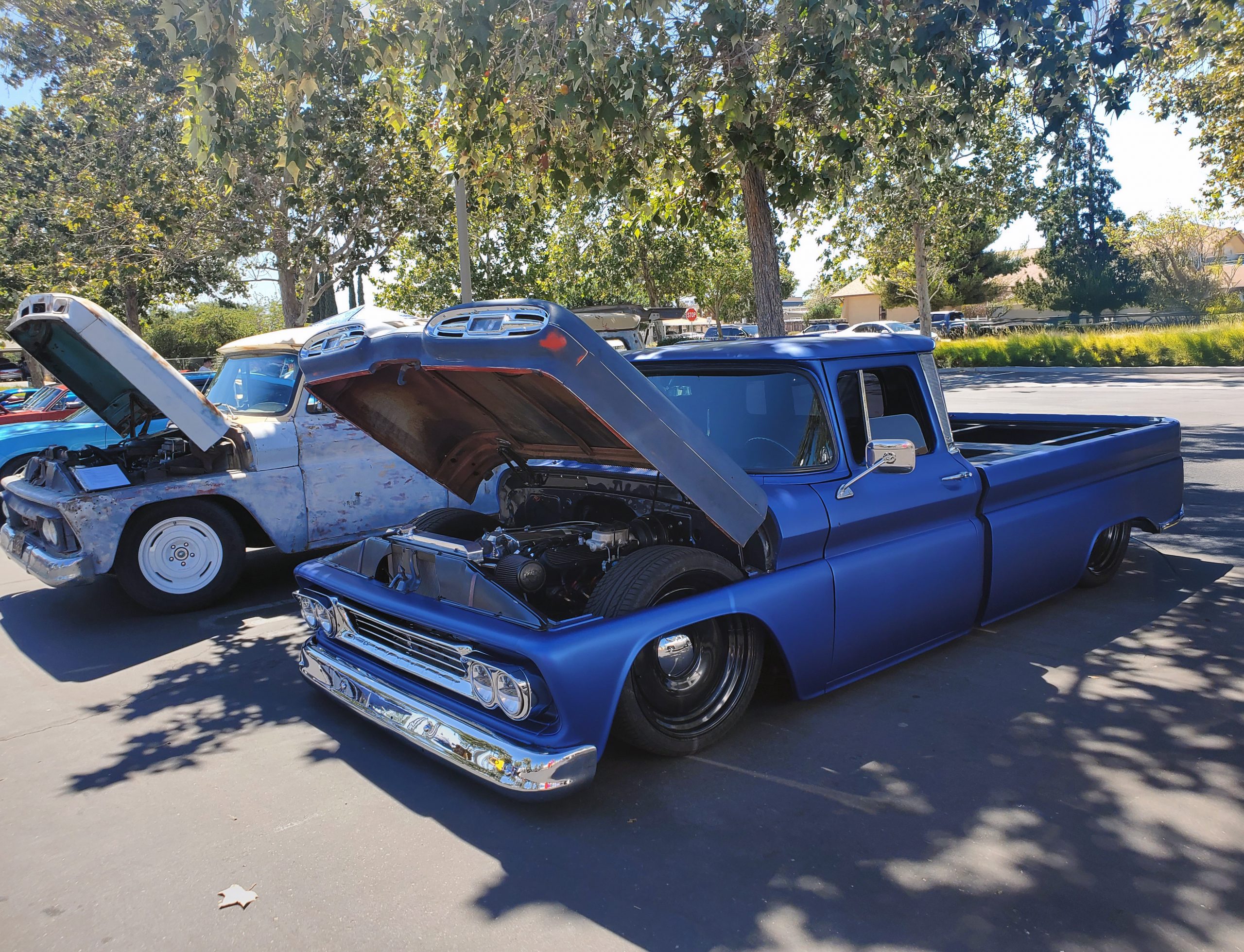 A bright coral vintage hot rod with its hood open, displaying a polished chrome engine, parked at an outdoor car show with other classic vehicles in the background.