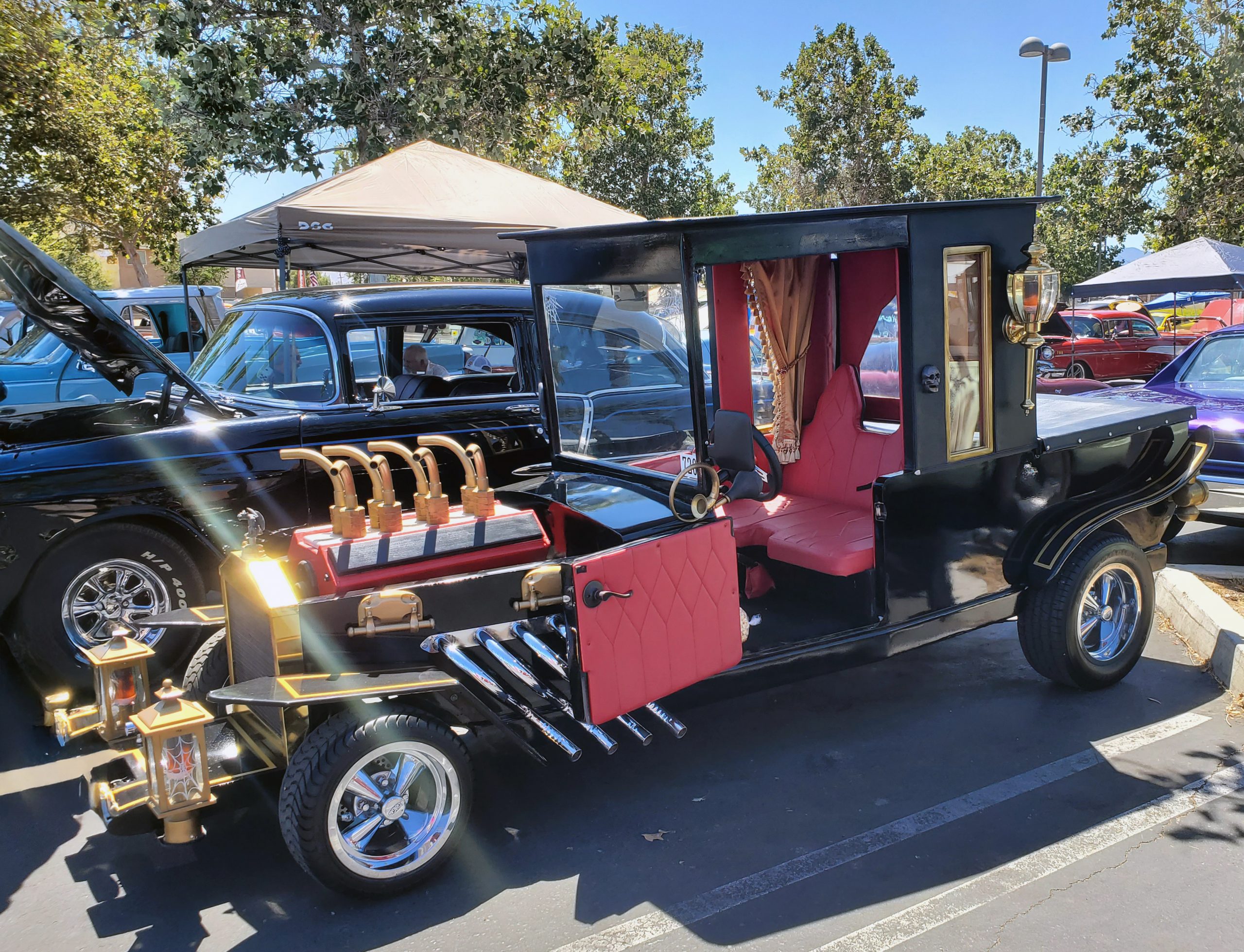 Vintage black steampunk-style car with brass fixtures, red leather interior, and exposed pipes displayed at a sunny outdoor auto show.