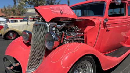 A bright coral vintage hot rod with its hood open, displaying a polished chrome engine, parked at an outdoor car show with other classic vehicles in the background.