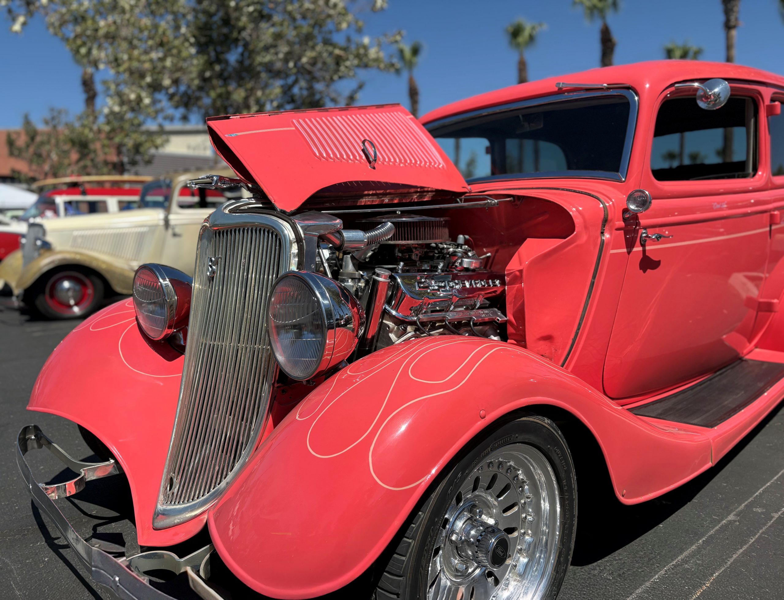 A bright coral vintage hot rod with its hood open, displaying a polished chrome engine, parked at an outdoor car show with other classic vehicles in the background.