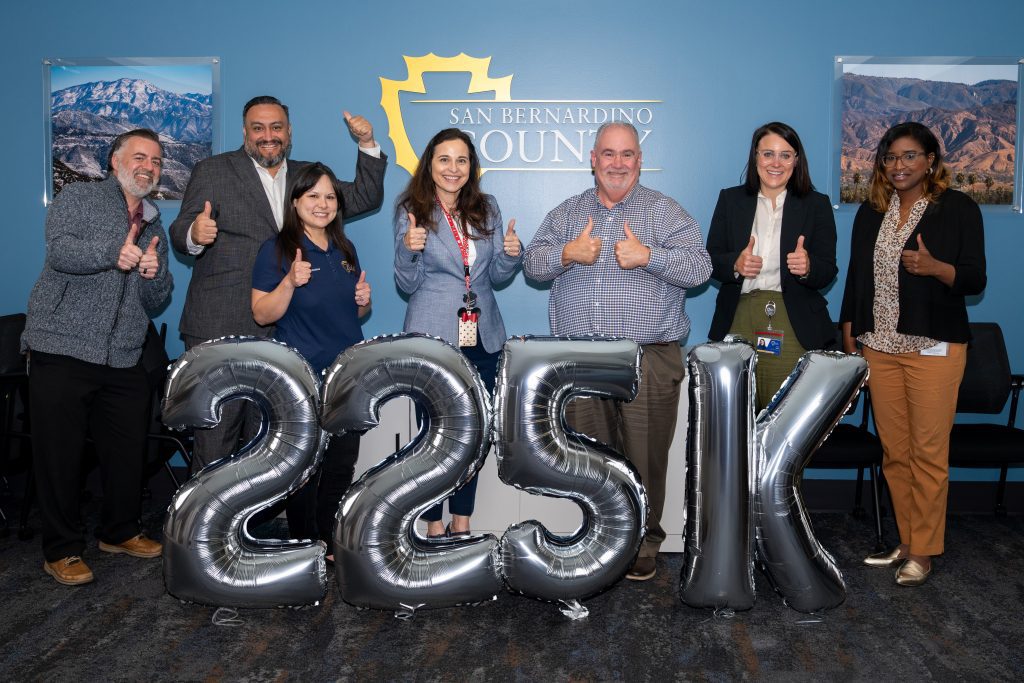 A group of people standing inside a room are smiling and giving thumbs up. There are large silver balloons that form the text "225K" in front of them.