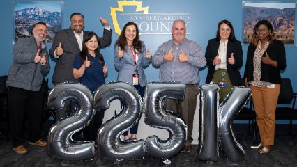 A group of people standing inside a room are smiling and giving thumbs up. There are large silver balloons that form the text "225K" in front of them.