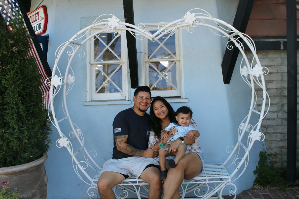 A smiling man and woman sit with a young child on a decorative heart-shaped bench outside a building.