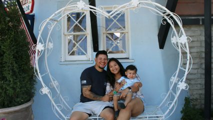 A smiling man and woman sit with a young child on a decorative heart-shaped bench outside a building.