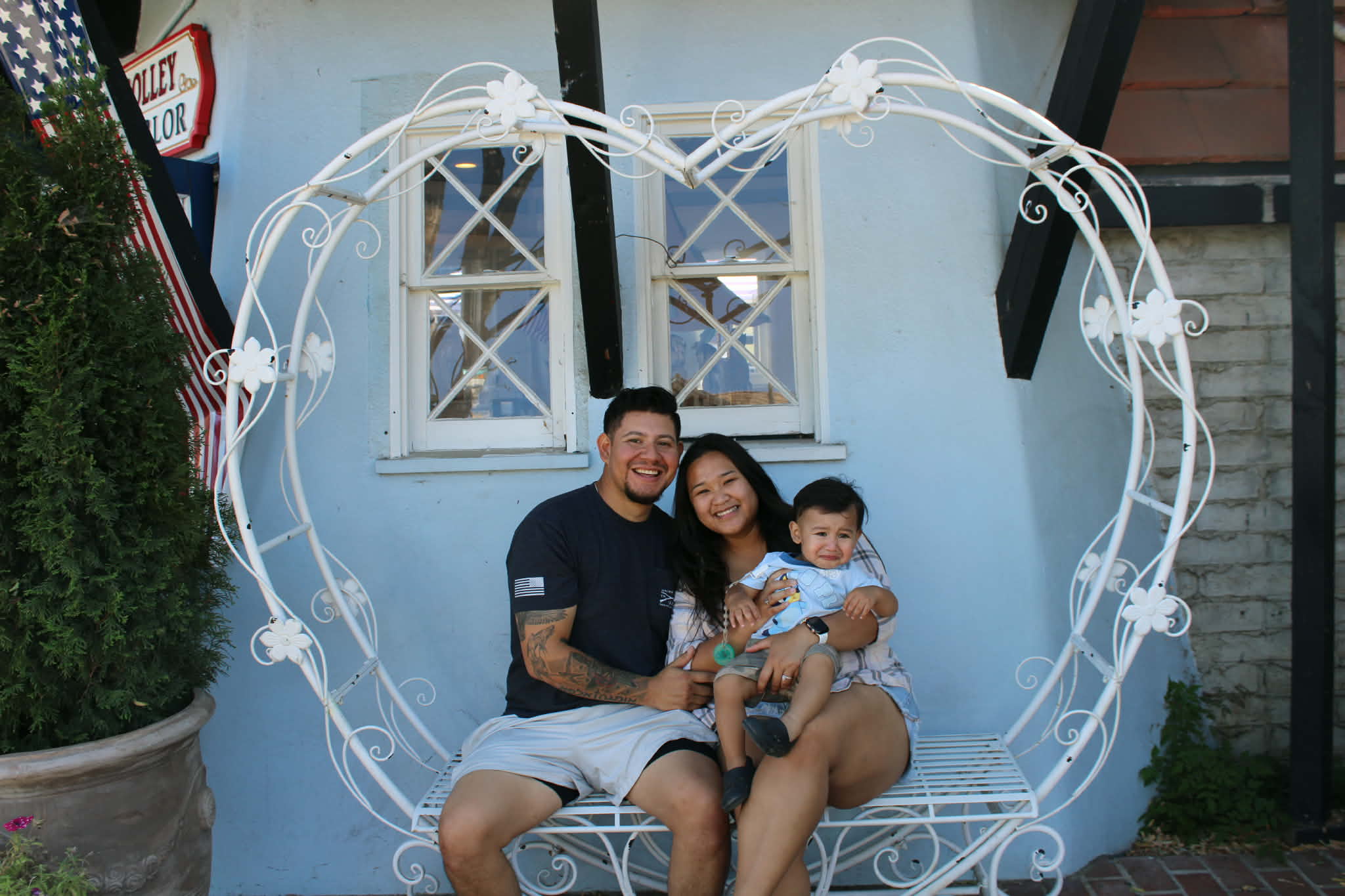 A smiling man and woman sit with a young child on a decorative heart-shaped bench outside a building.