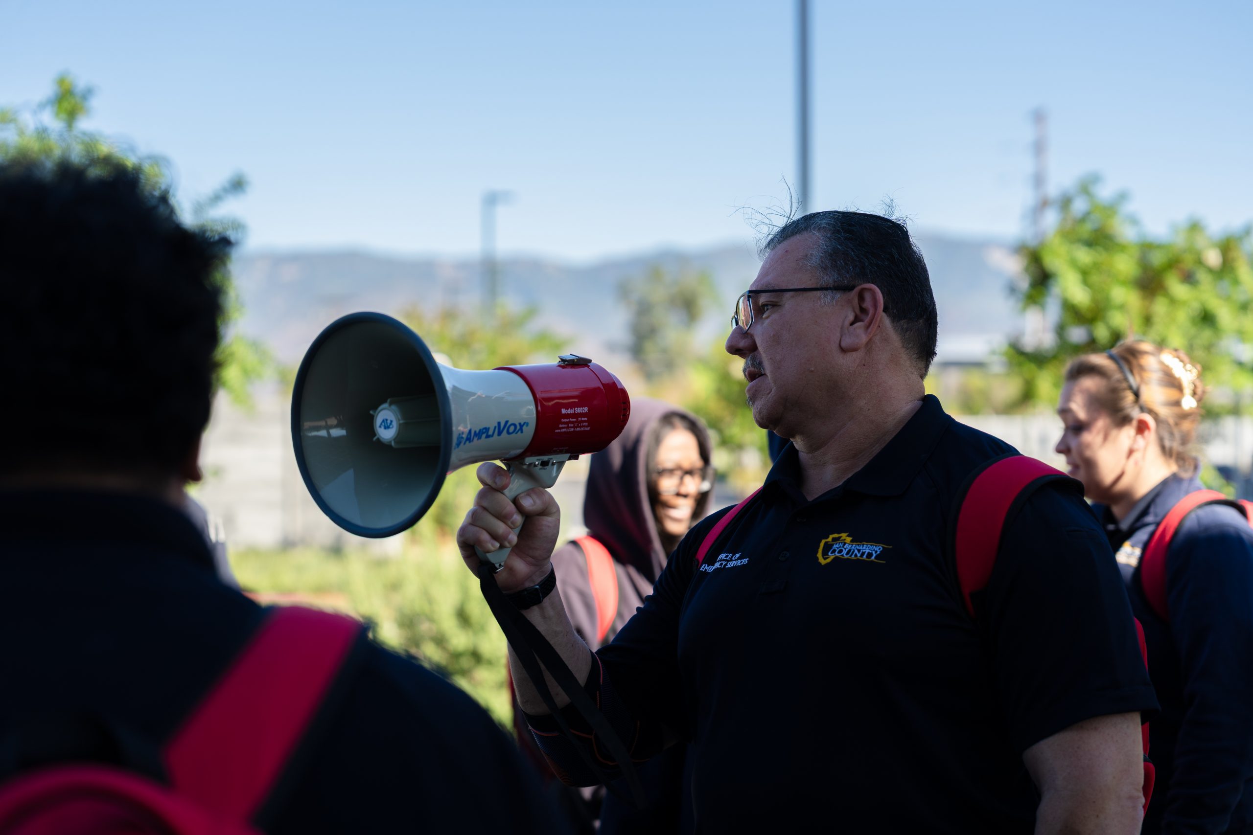A man speaks into a mega phone outside, wearing a uniform and red back pack as other employees surround him wearing uniforms and red backpacks.