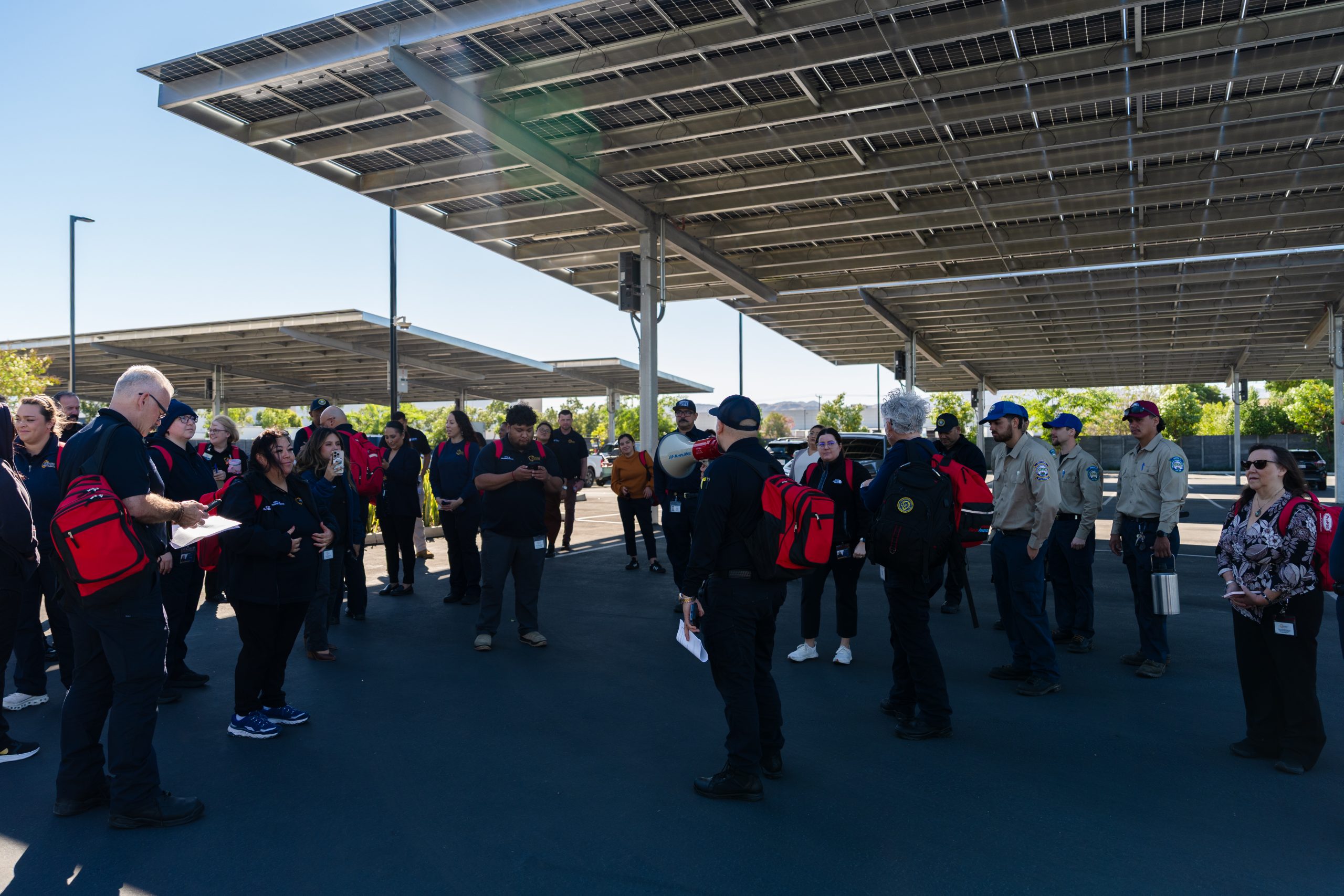 A large group of employees in uniform assemble under solar panels in a parking lot.