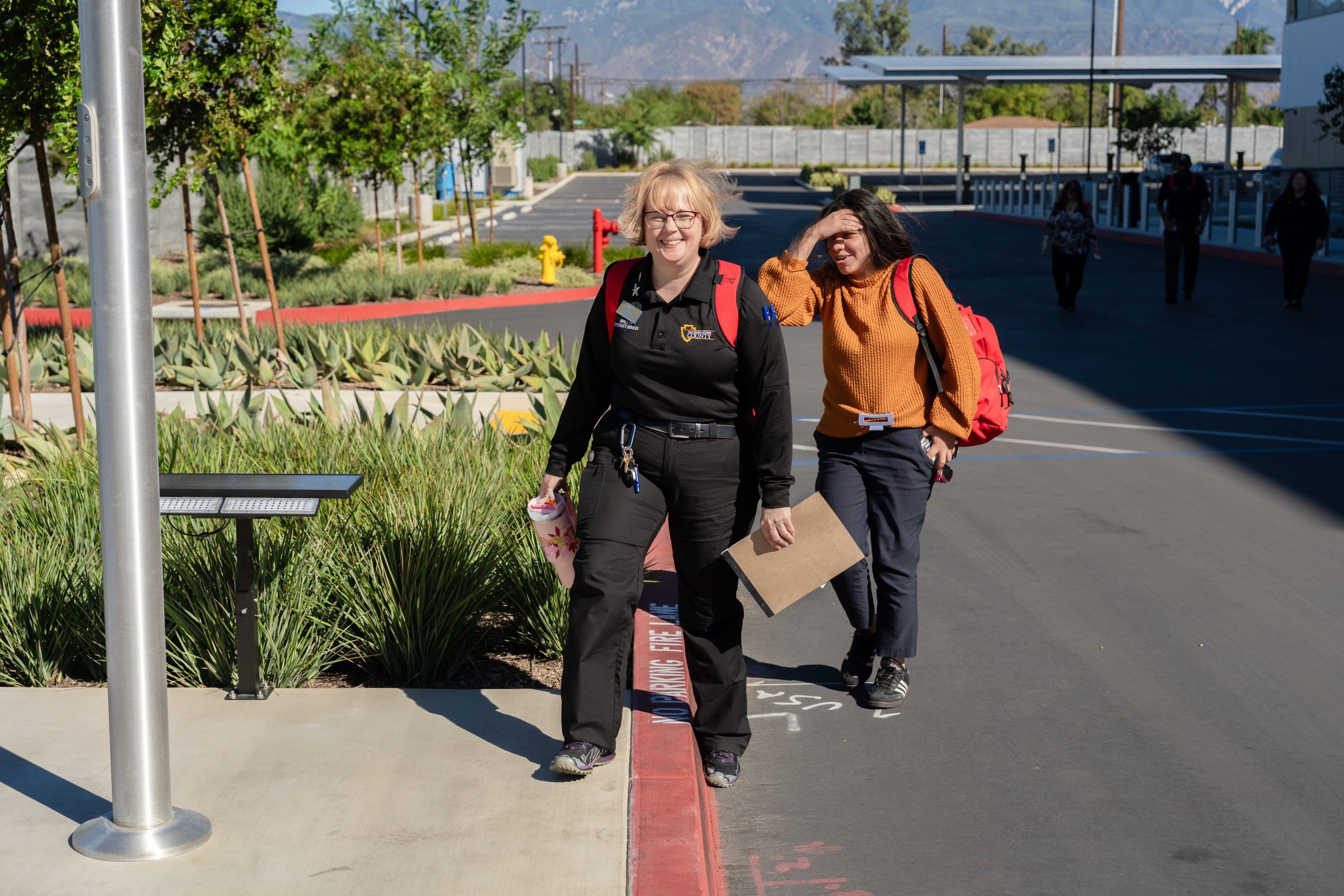 Two women walk onto a sidewalk in a parking lot, wearing red backpacks, on a sunny day.
