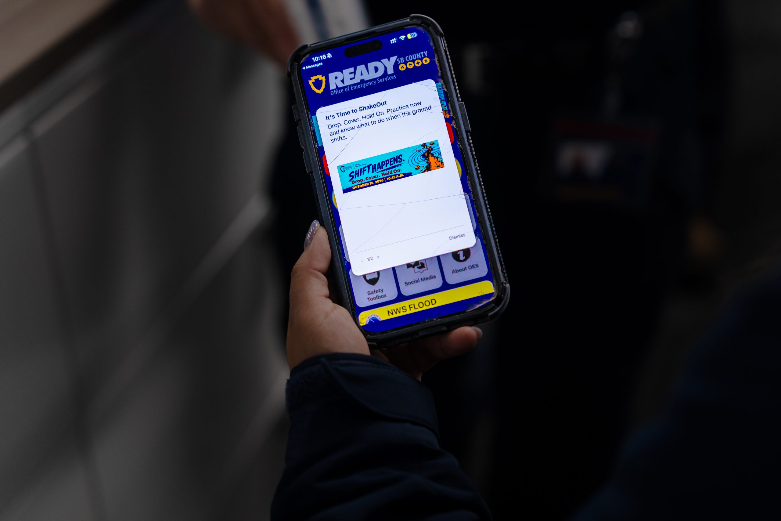 A person holds a phone with the "Ready" SB County app displayed, showing the "Shift Happens" banner for the Great ShakeOut drill and exercise.