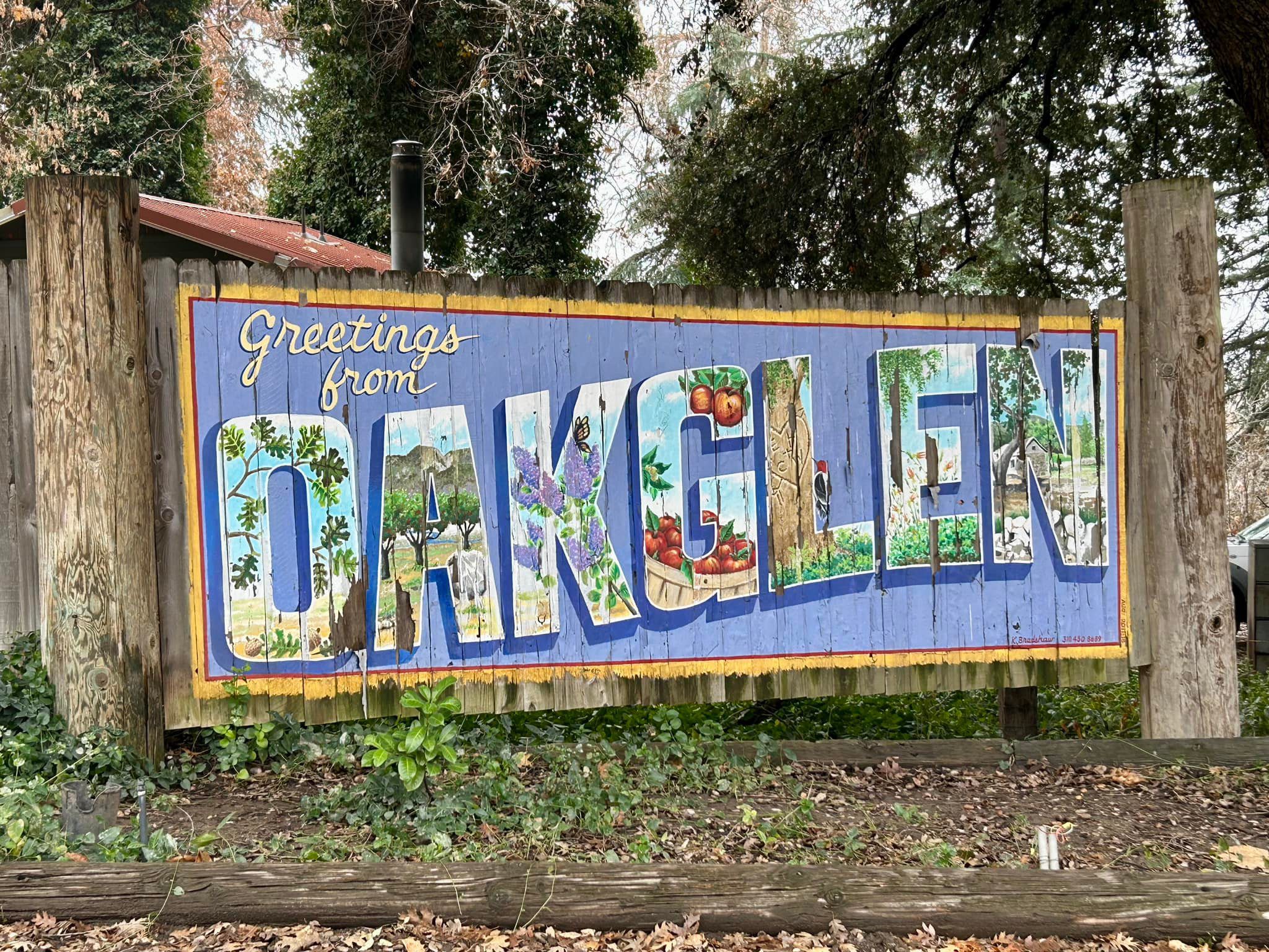 A weathered illustrated sign welcoming visitors to Oak Glen, with each letter of 'OAK GLEN' showcasing different aspects of the region, such as apple baskets, trees, and mountain landscapes. The sign is nailed to a wooden fence with plants and trees growing around it."