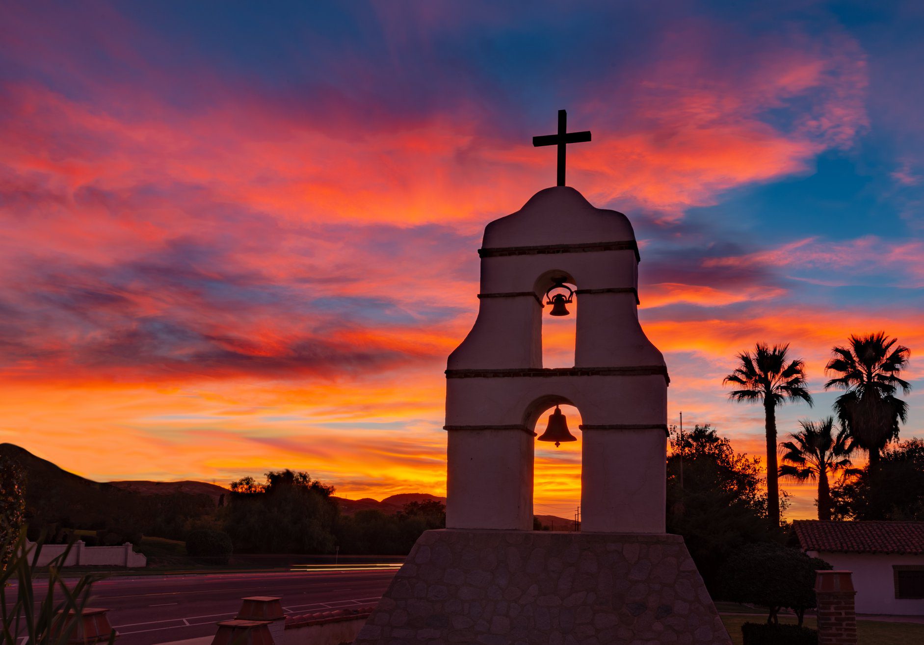 An image of a bell tower with a cross on top taken at sunset featuring palm trees and hills in the background.