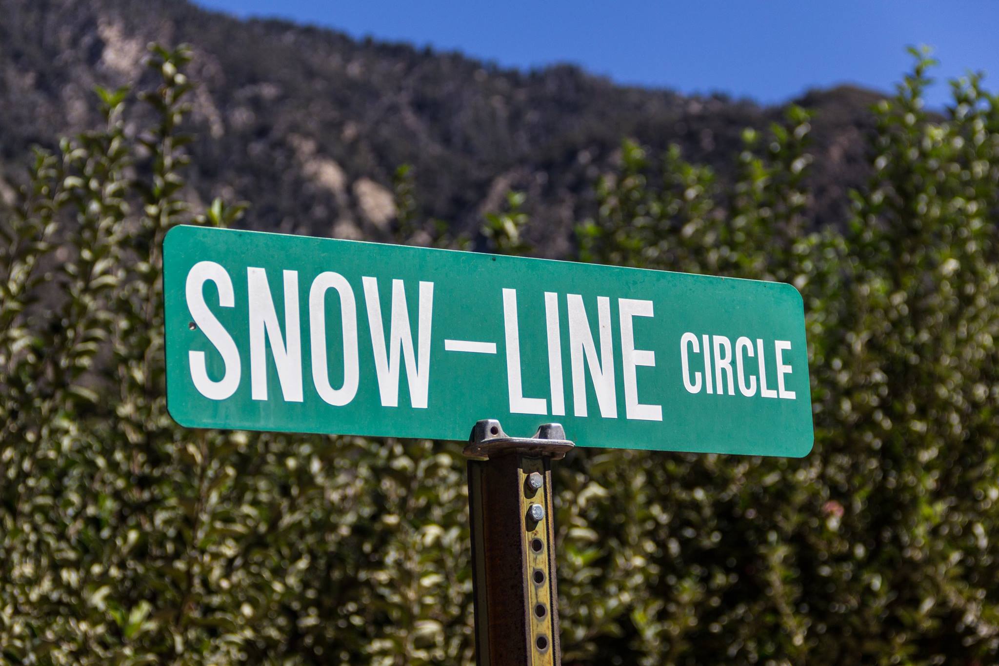 A street sign for 'SNOW-LINE CIRCLE' in Oak Glen, standing against a backdrop of green foliage, mountain terrain, and a clear blue sky.