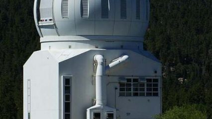 A large and white observatory dome near a forested area and lake.