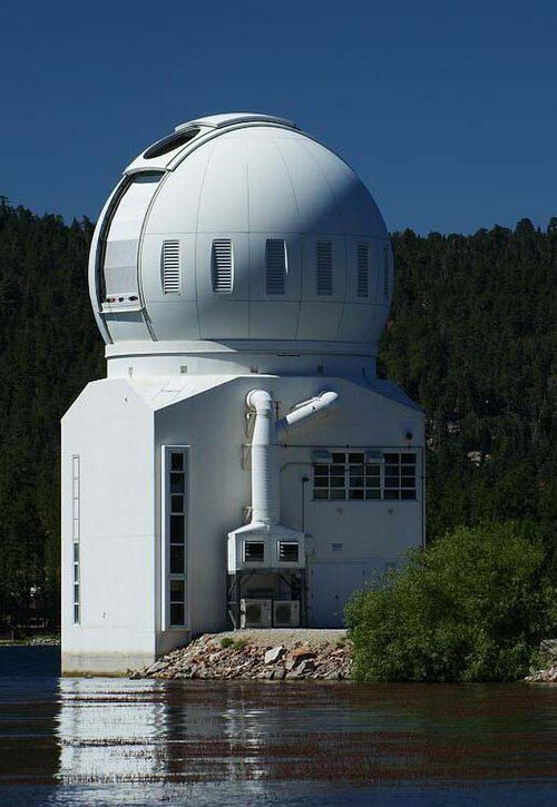 A large and white observatory dome near a forested area and lake.