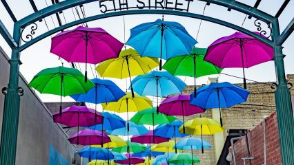 Pink, blue, yellow, and green umbrellas are hanging above a walkway near a metal arch labeled "5th Street."