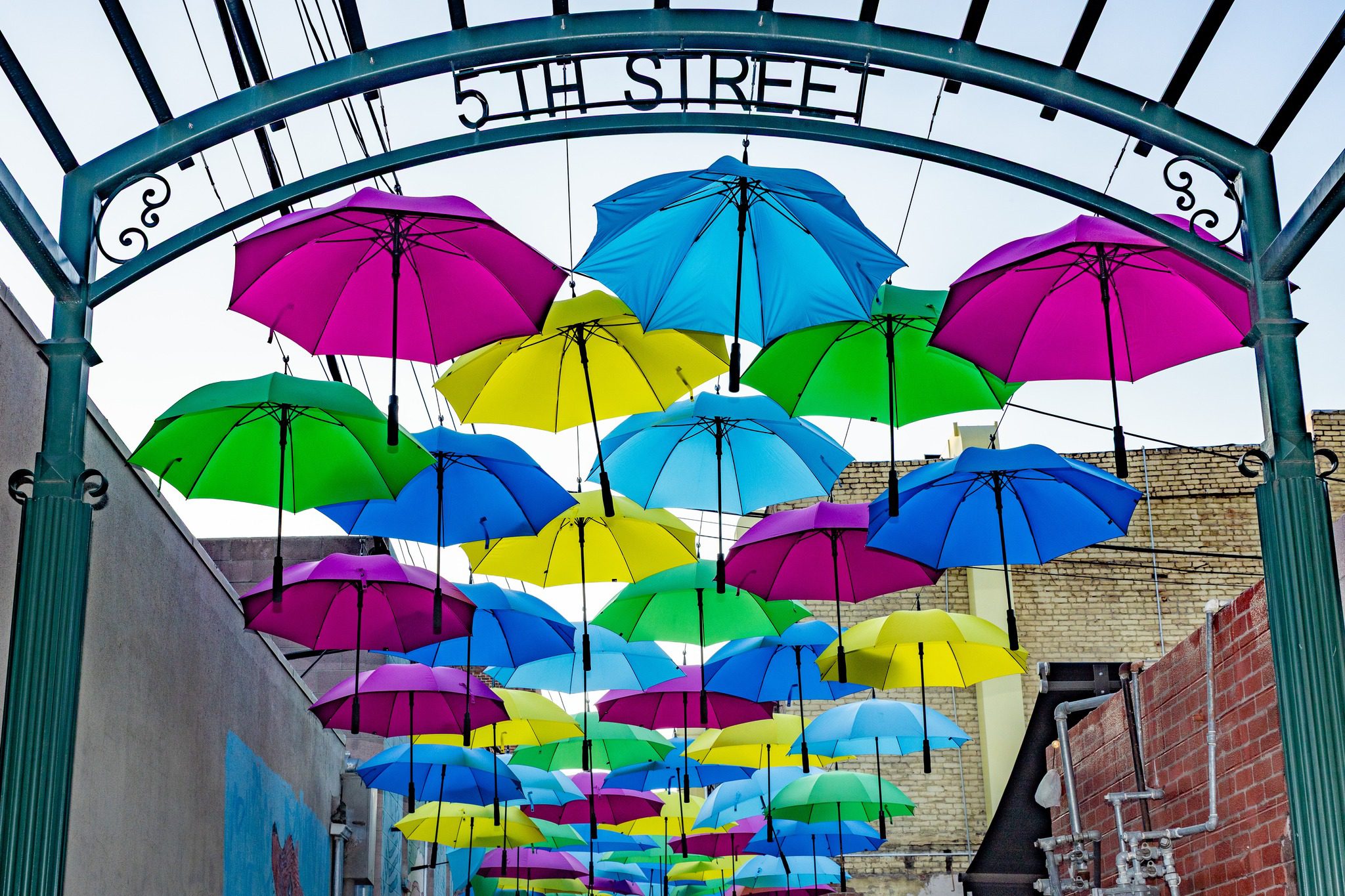 Pink, blue, yellow, and green umbrellas are hanging above a walkway near a metal arch labeled "5th Street."