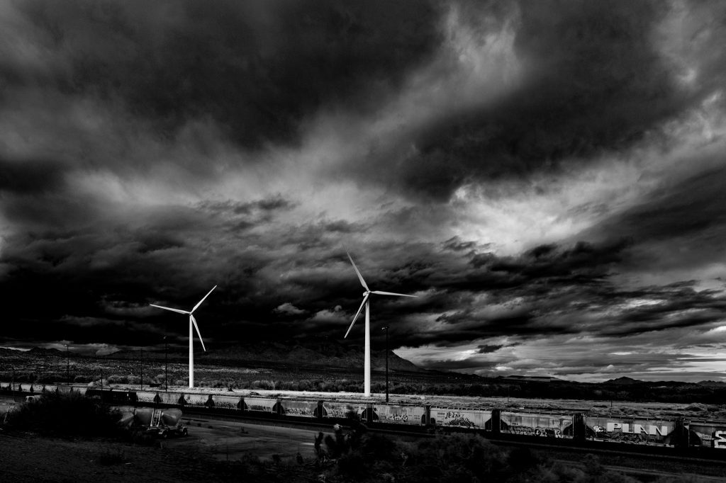 A black and white image of a freight train with windmills in the background. The sky is filled with dark and swirling clouds.