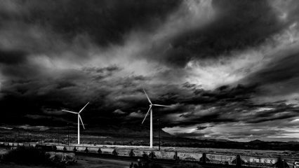 A black and white image of a freight train with windmills in the background. The sky is filled with dark and swirling clouds.